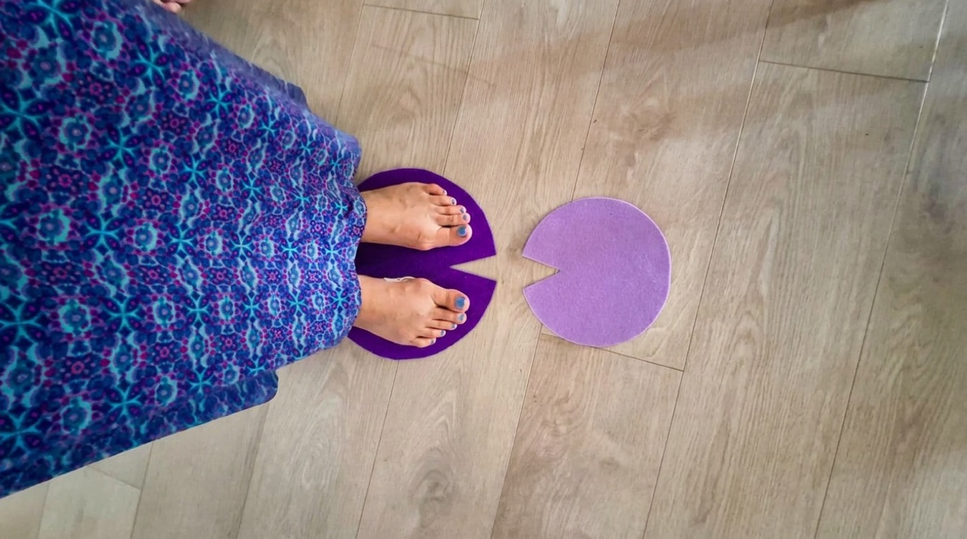 Bare feet standing on a purple felt marker cut in a curved shape, with a second lilac marker placed apart on a wooden floor.